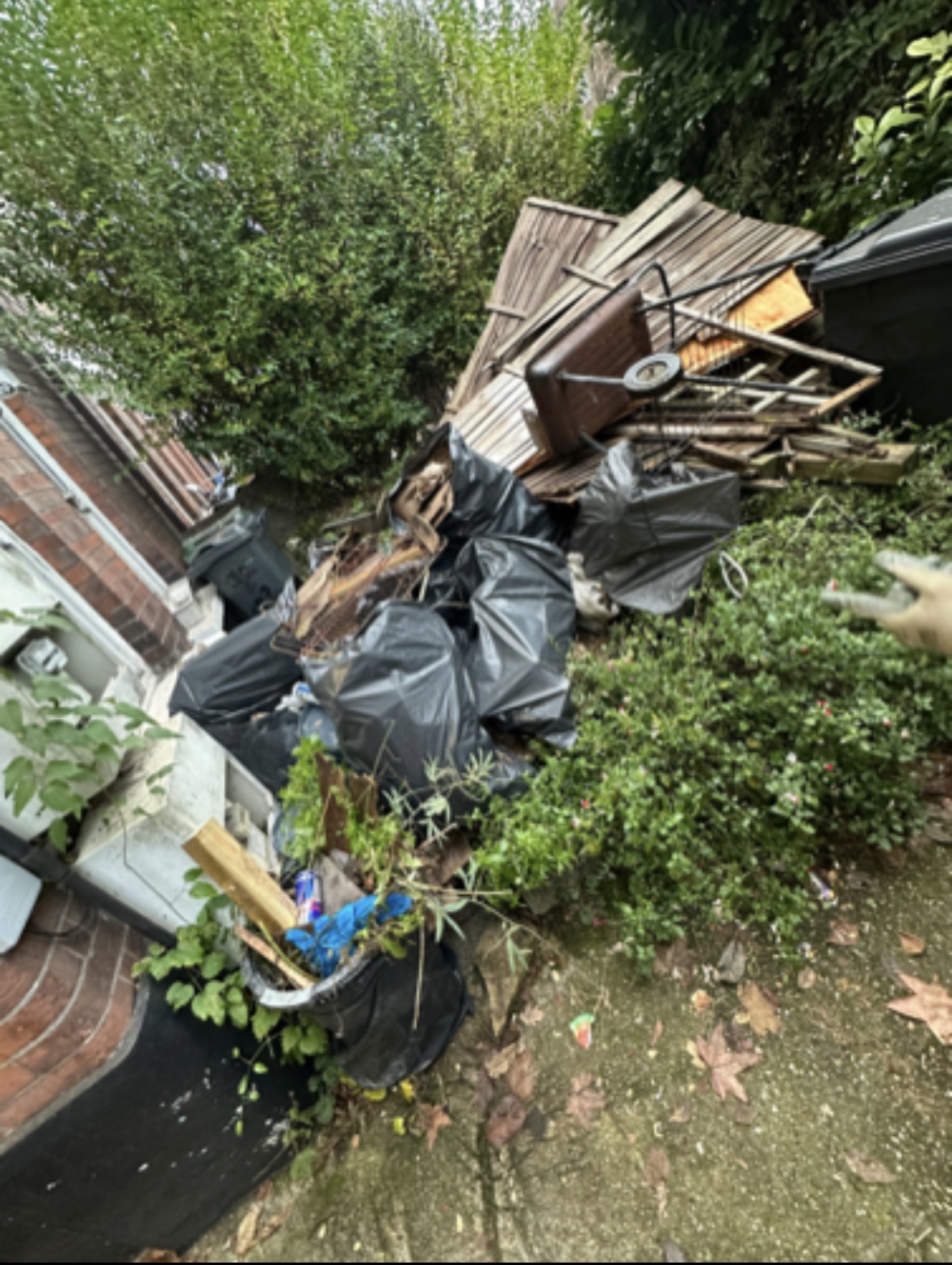 Mixed garden waste, sacks and broken fencing stacked beside a house.